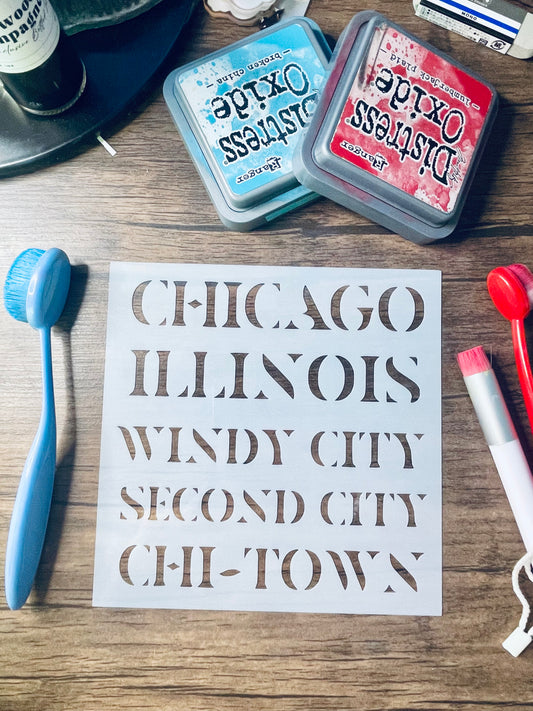 A white stencil sheet with the words 'CHICAGO ILLINOIS WINDY CITY SECOND CITY CHI-TOWN' laid on a wooden surface, surrounded by craft tools and two Distress Oxide ink pads.
