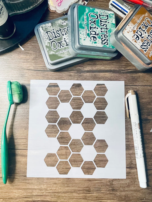 A white square stencil sheet with a repeating hexagon pattern laid on a wooden surface, surrounded by craft tools including a green-handled brush, a white marker, and three Distress Oxide ink pads.