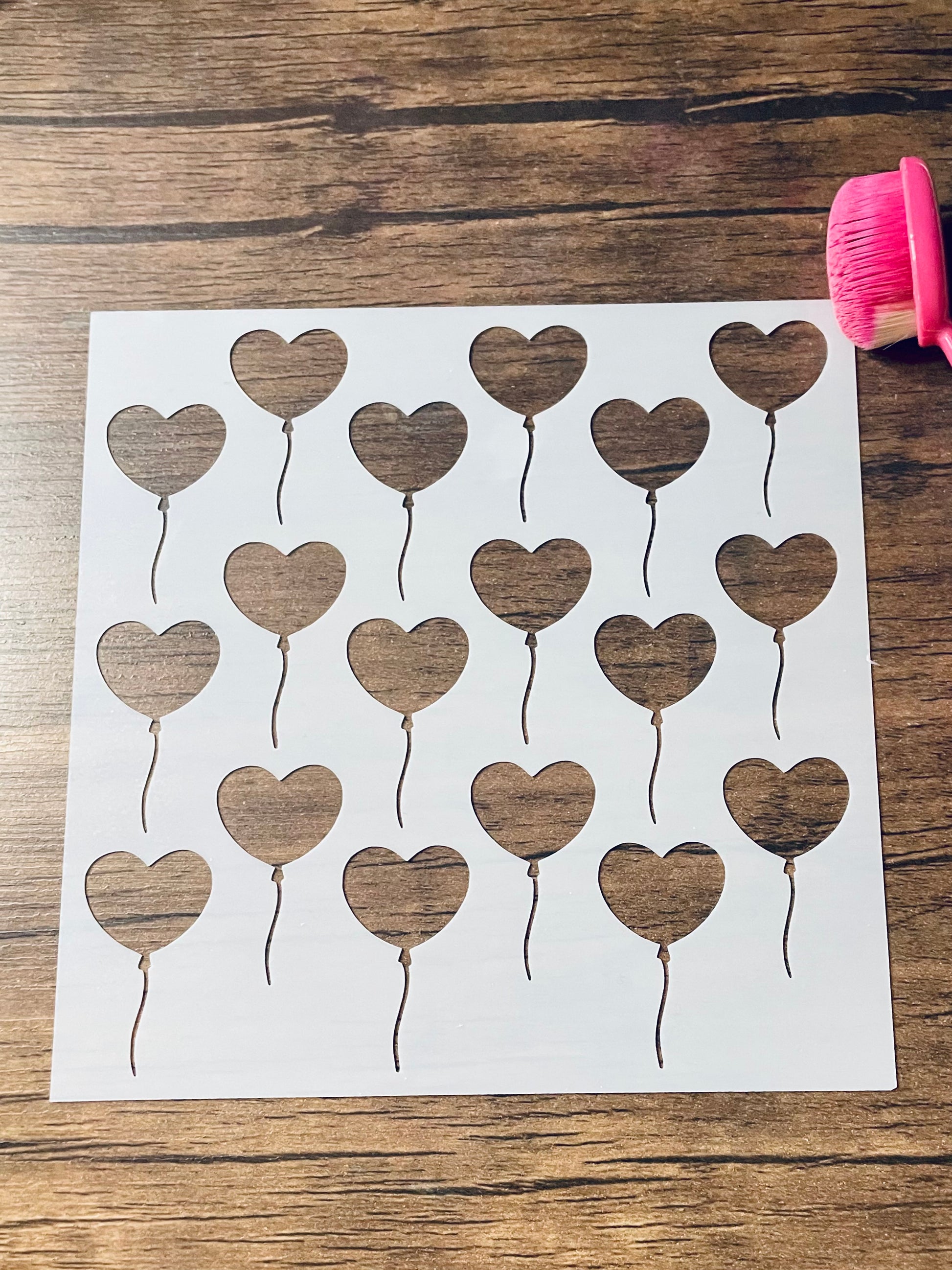 5.75” x 5.75” frosted stencil with Silhouettes of: 18 heart shaped balloons in three rows in a roughly diagonal pattern.  Stencil is on a wood surface with a pink brush 
