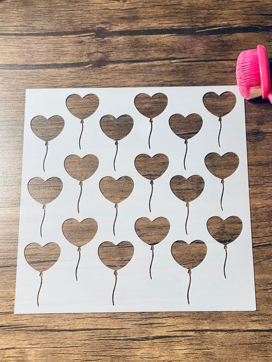 5.75” x 5.75” frosted stencil with Silhouettes of: 18 heart shaped balloons in three rows in a roughly diagonal pattern.  Stencil is on a wood surface with a pink brush 