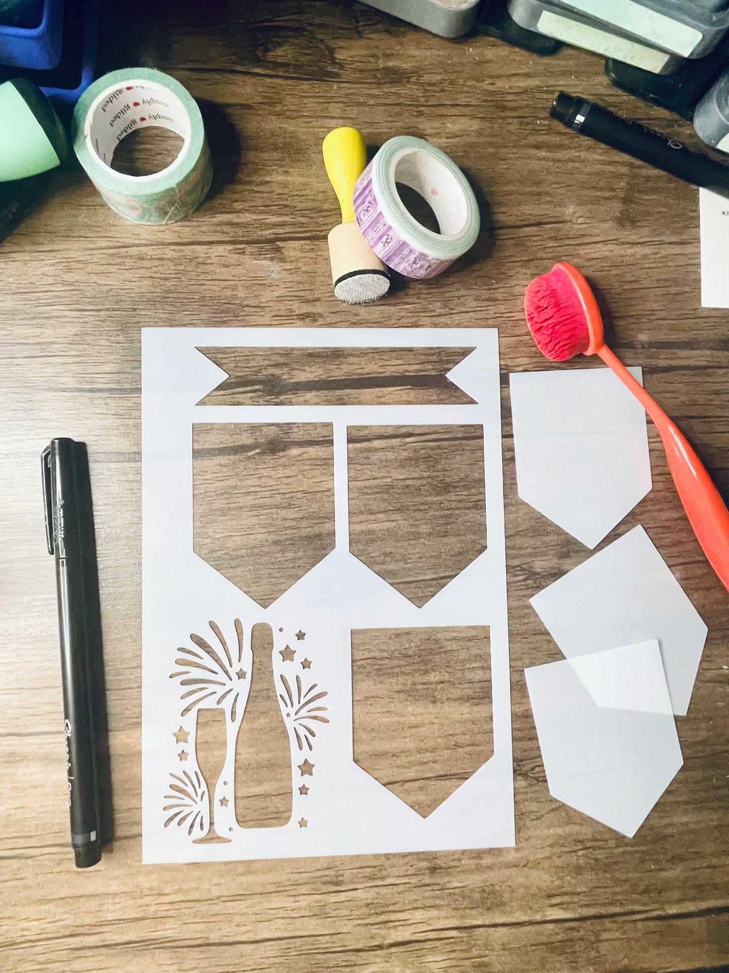 a rectangular stencil sits on a wooden desk.  Also on the desk is a red brush and other stationary.  The stencil is frosted and includes cutouts of:  a banner on top, a row of 2 pentagon shaped banners, followed by one more on the bottom. next to bottom banner is a champagne bottle, champagne flute, and fireworks and stars behind them.  The cutouts of the 3 pentagon shaped stencils are popped out and to the right of the stencil
