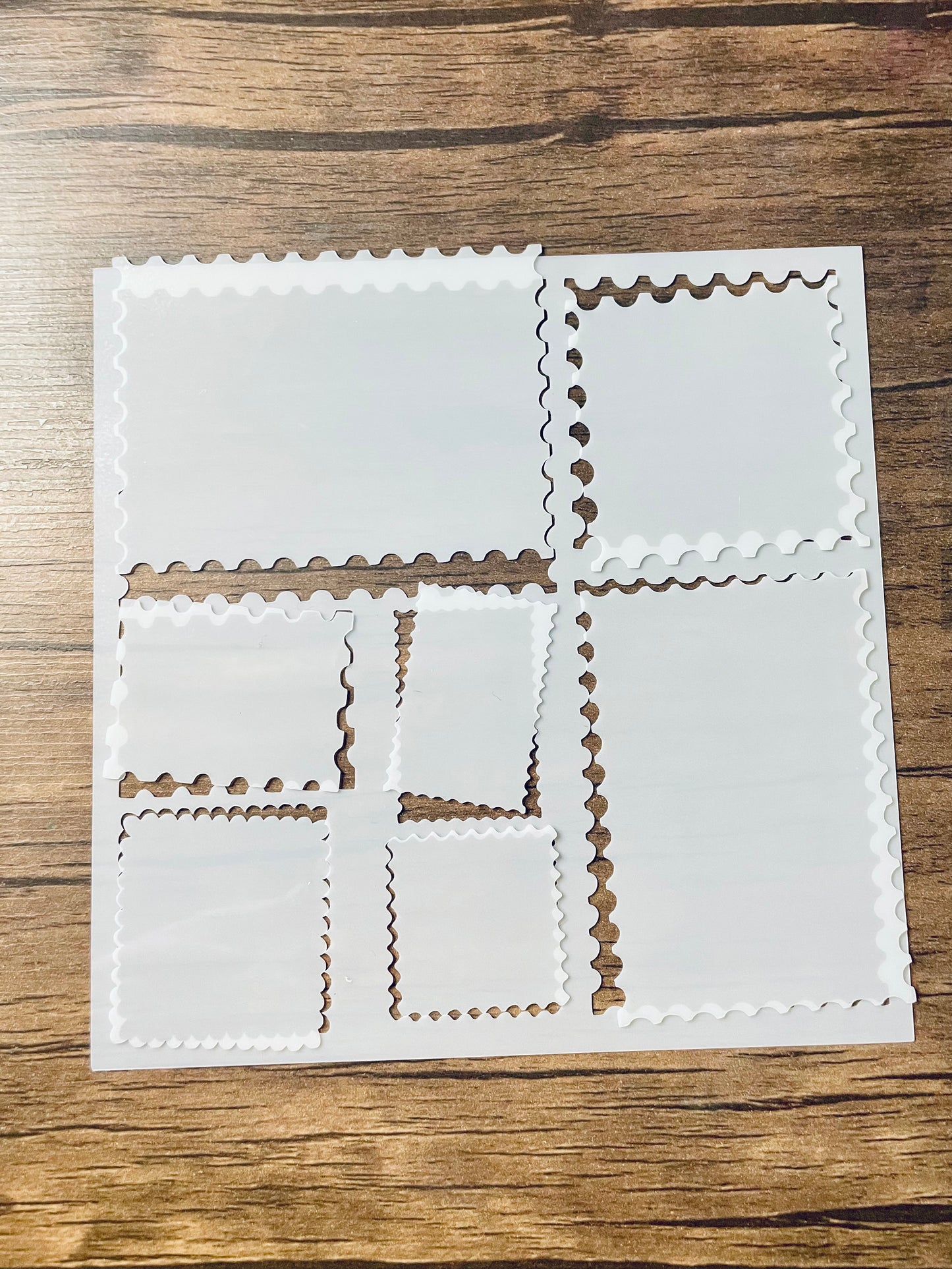 5.75” x 5.75” frosted stencil with Silhouettes of: 7 different stamp shaped cutouts.  The cutout pieces are loosely rested in their cut out shape.  Stencil is on a wood surface with a pink brush 