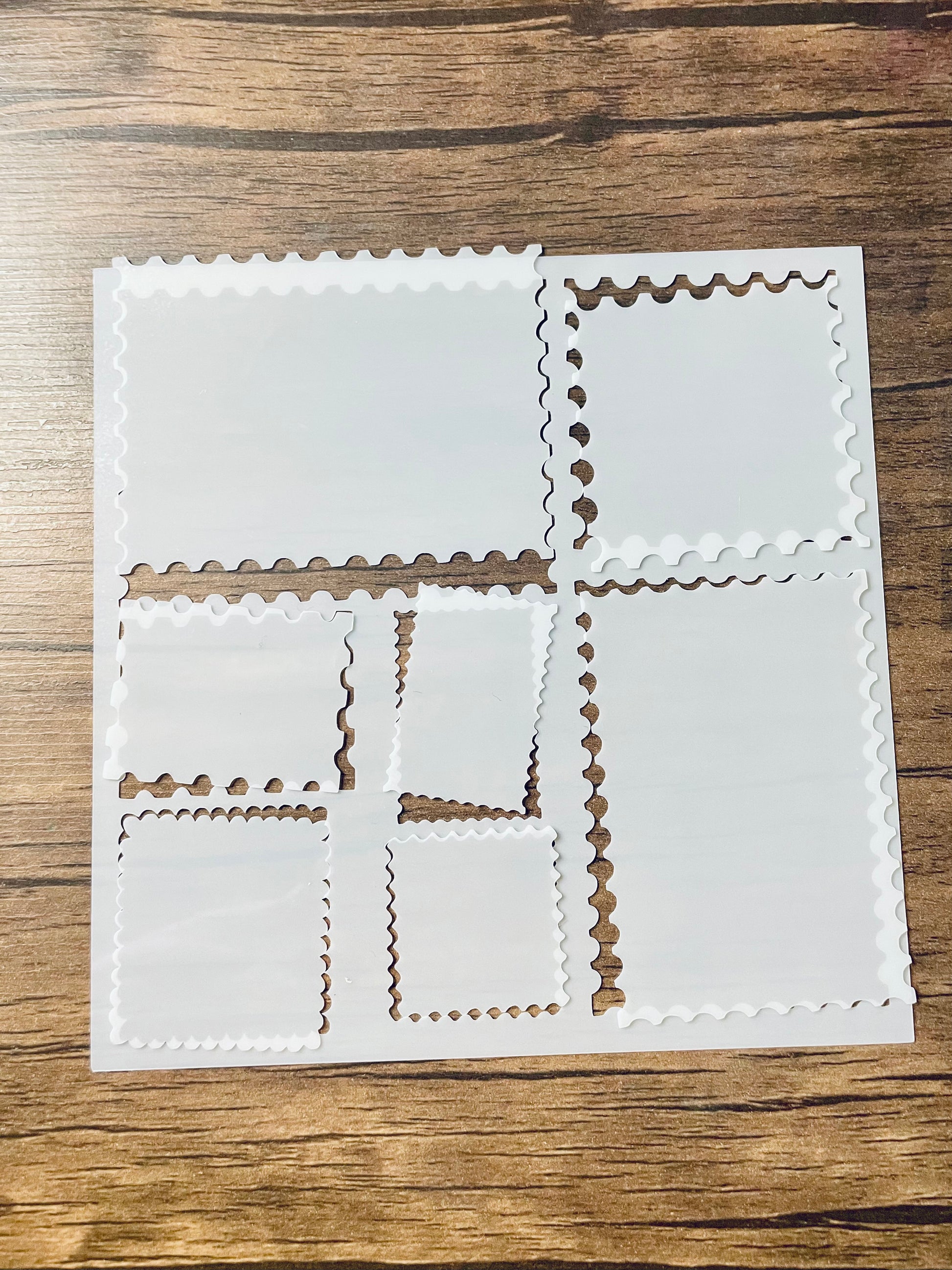 5.75” x 5.75” frosted stencil with Silhouettes of: 7 different stamp shaped cutouts.  The cutout pieces are loosely rested in their cut out shape.  Stencil is on a wood surface with a pink brush 