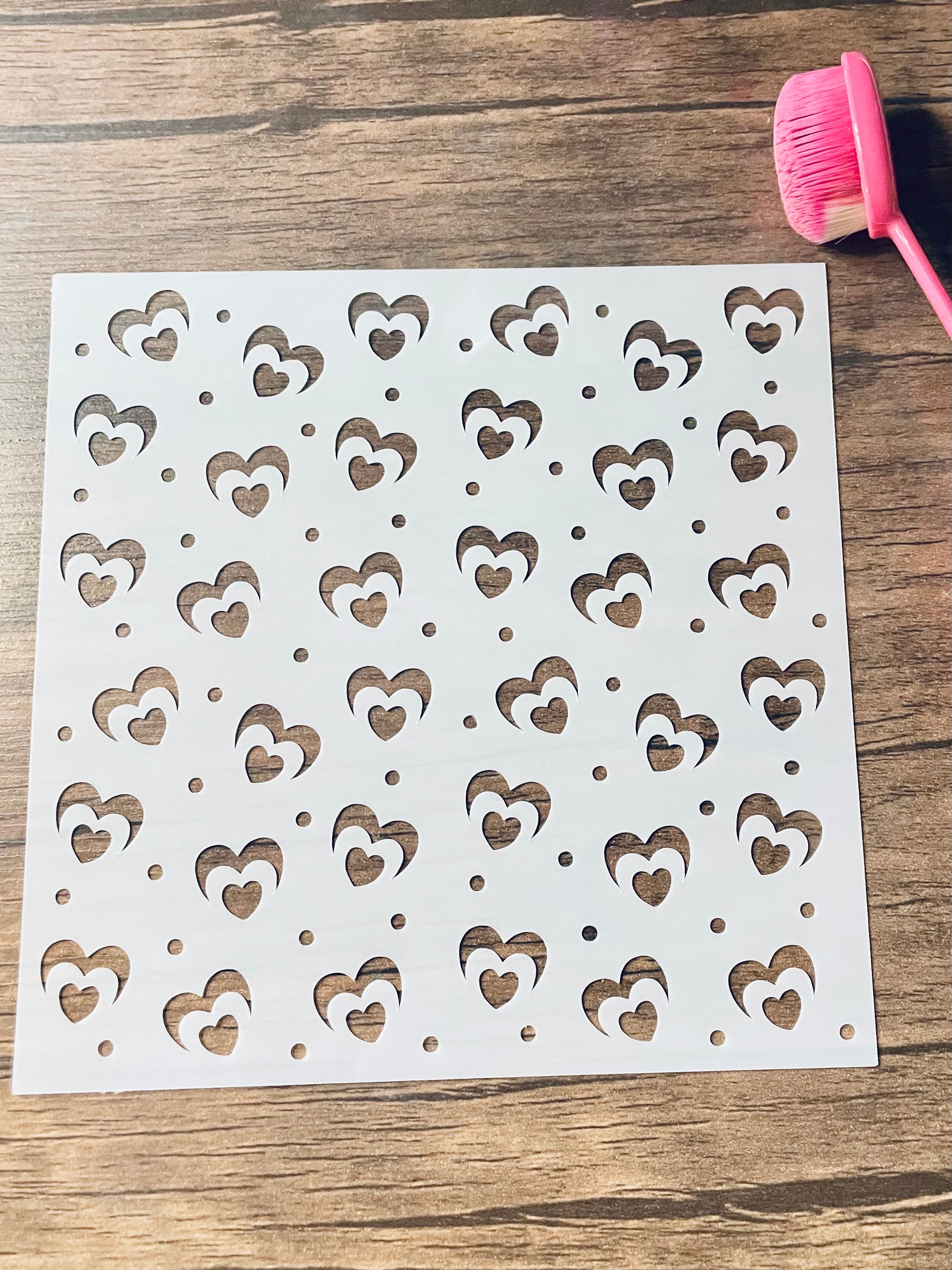5.75” x 5.75” frosted stencil with Silhouettes of: Hearts with a booming heart in a pattern around the stencil on a wooden surface with a pink brush.