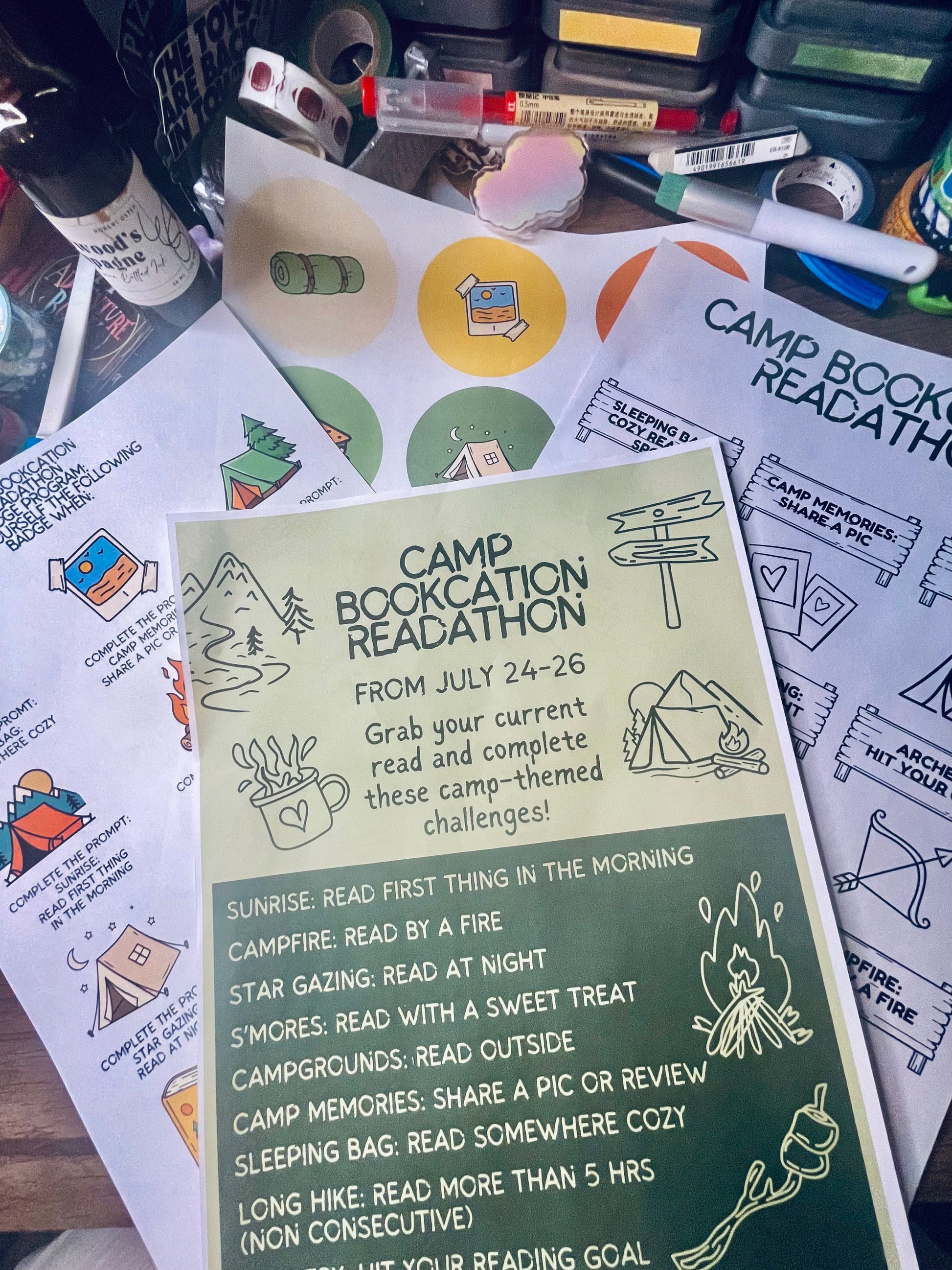 A printed 'Camp Book Club Readathon' activity sheet laid on top of other camping-themed printed cards and worksheets on a cluttered desk. The main sheet has a pale yellow top half with black text and illustrations, and a dark green bottom half with reading goals and activity prompts.