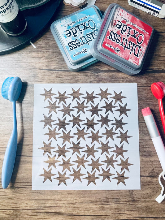 A sheet of white stencil paper with rows of brown star cutouts, laid on a wooden surface with craft tools and Tim Holtz Distress Oxide ink pads around it.