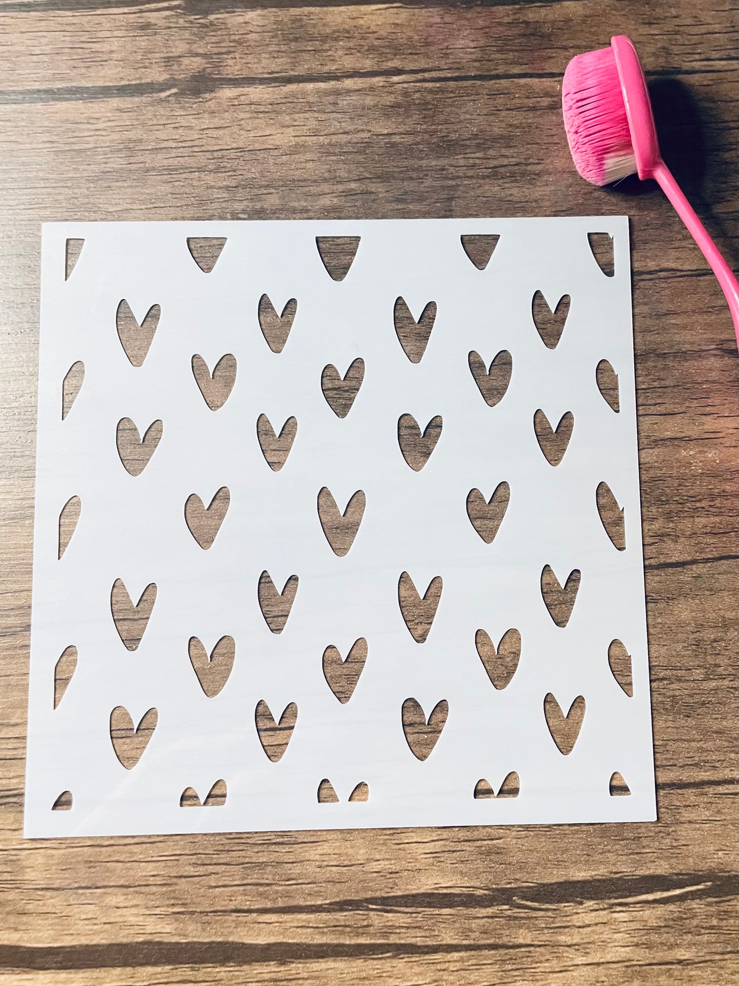 5.75” x 5.75” frosted stencil with Silhouettes of: small hearts in a doodles whimsical version in a pattern .  Stencil is on a wood surface with a pink brush 