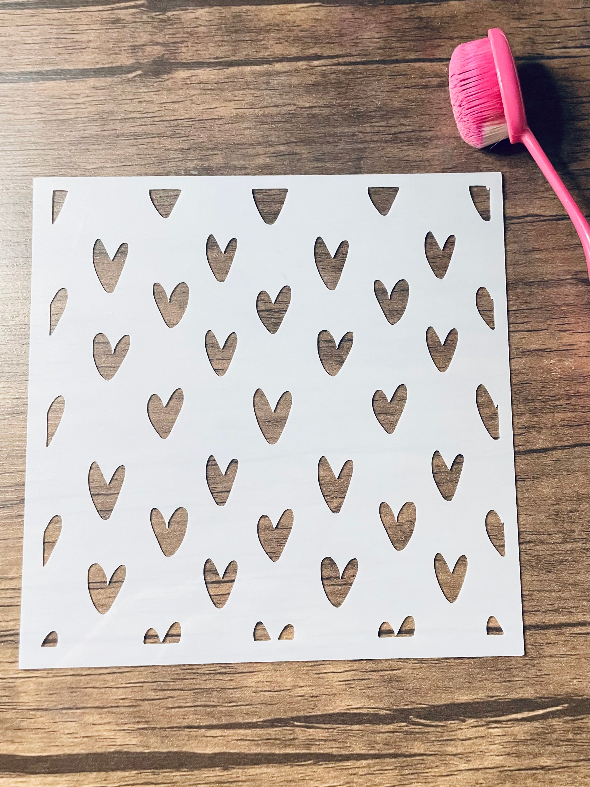 5.75” x 5.75” frosted stencil with Silhouettes of: small hearts in a doodles whimsical version in a pattern .  Stencil is on a wood surface with a pink brush 