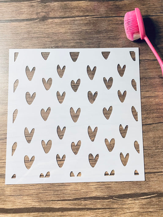 5.75” x 5.75” frosted stencil with Silhouettes of: small hearts in a doodles whimsical version in a pattern .  Stencil is on a wood surface with a pink brush 