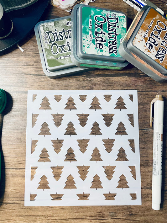 A square white card with a repeating pattern of brown Christmas tree silhouettes, laid on a wooden desk. Three Distress Oxi ink pads in green, blue, and bronze are stacked at the top, with a white pen at the right.