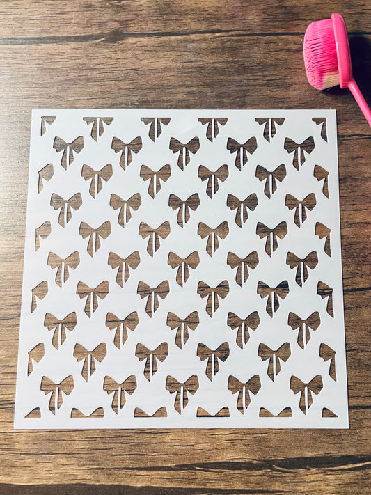 5.75” x 5.75” frosted stencil with Silhouettes of: small bows in a roughly diagonal pattern .  Stencil is on a wood surface with a pink brush 