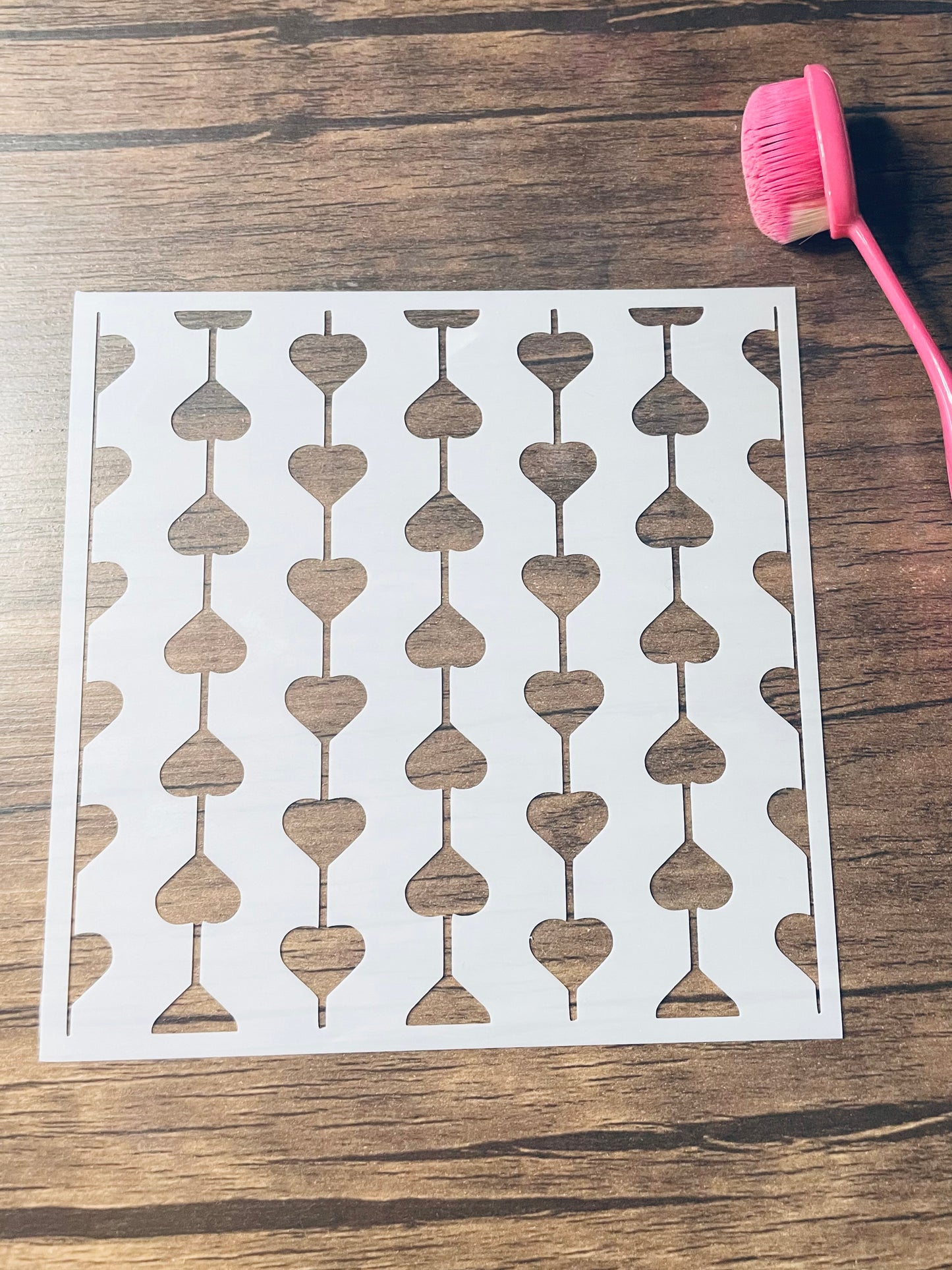 5.75” x 5.75” frosted stencil with Silhouettes of: hearts vertically strung together.   Each string alternates direction.   Stencil is on a wood surface with a pink brush 