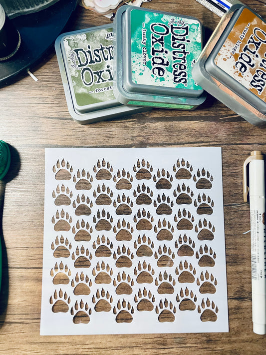 A white square stencil sheet with multiple paw print cutouts, placed on a wooden surface with Distress Oxi ink pads and a white marker nearby.