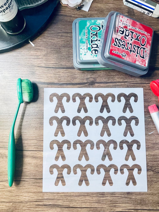 A sheet of white stickers printed with brown gingerbread man shapes, displayed on a wooden tabletop alongside a green paintbrush, Distress Oxide ink pads, and other craft supplies.