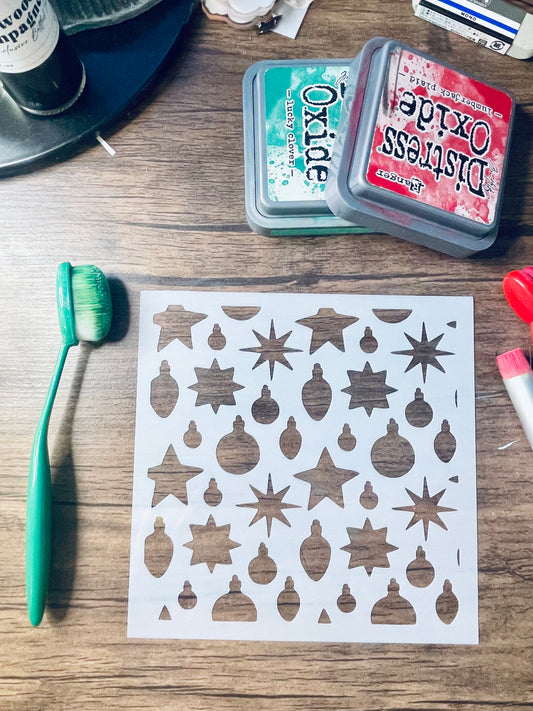 A sheet of white stencils with various star, starburst, and ornament shapes laid out on a wooden table, surrounded by craft supplies including an orange-handled brush, an Oxide Distress Ink pad, and other small tools.